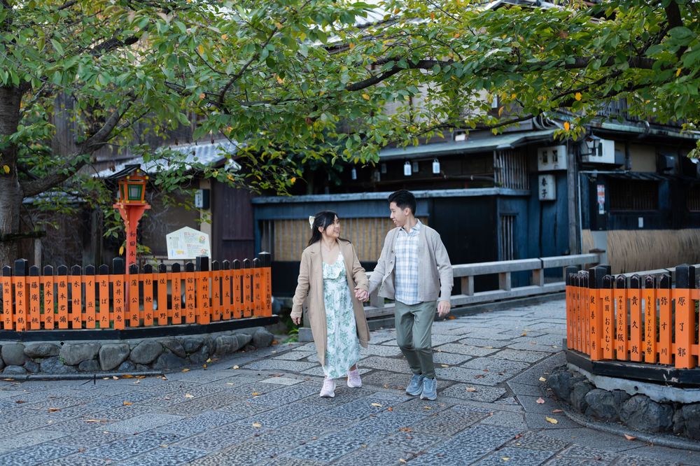 Couple holding hands strolling along a stone path beside traditional Japanese wooden buildings and orange railings under lush green trees.