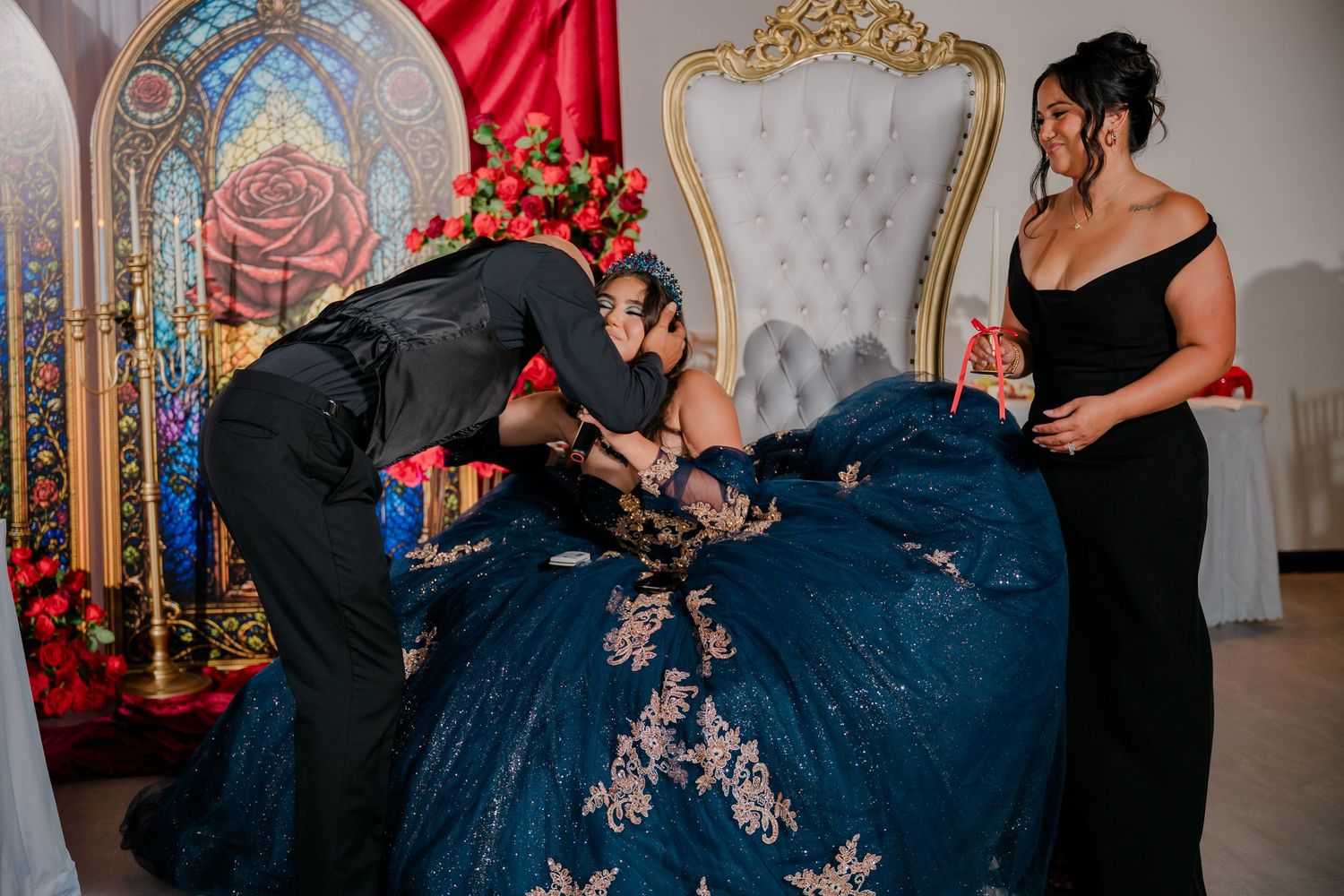 The father kneels and embraces a quinceañera in an embellished blue gown beside a gold throne chair as a woman in black watches.