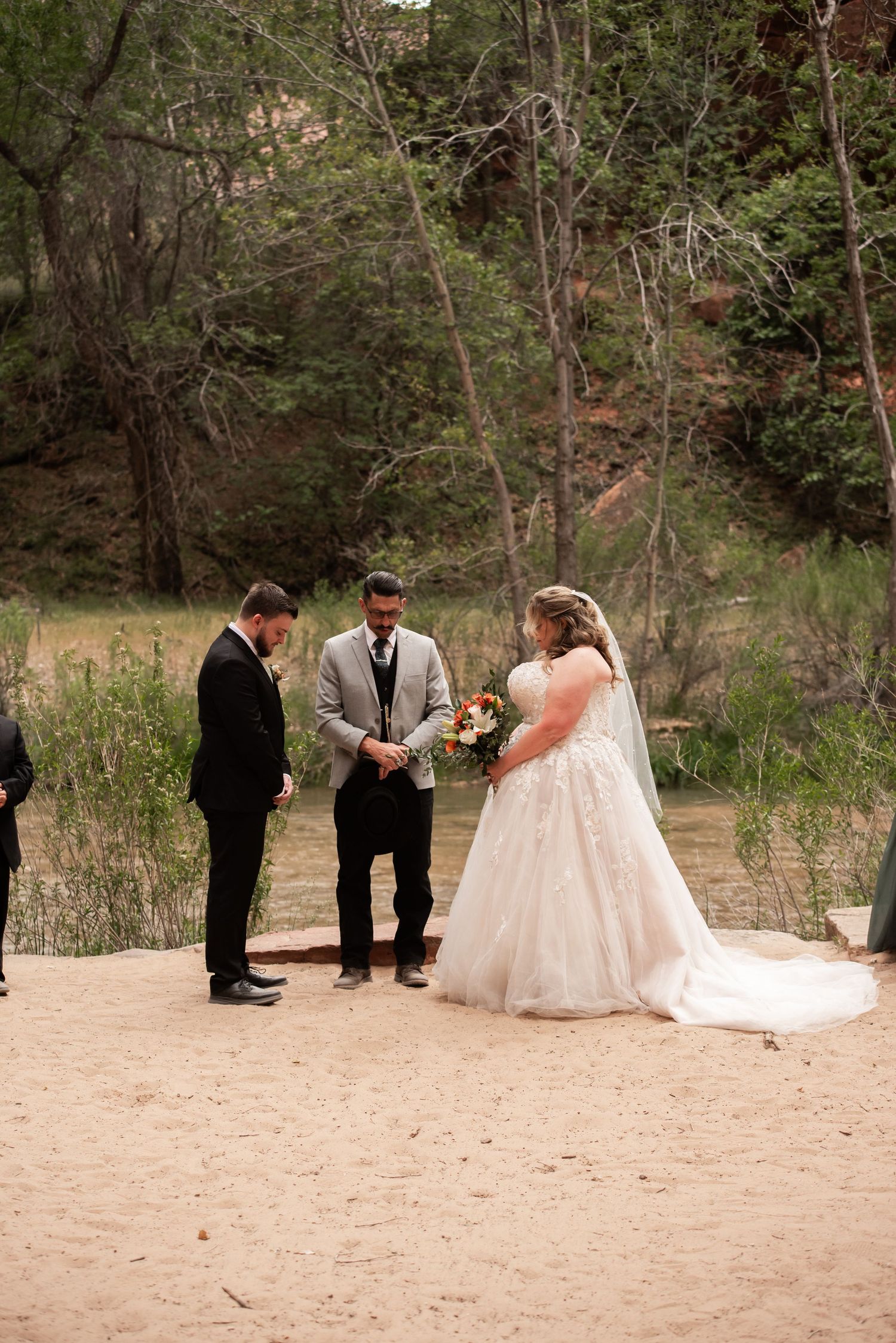 Outdoor wedding ceremony on sandy ground with bride in white gown and groom exchanging vows before officiant.
