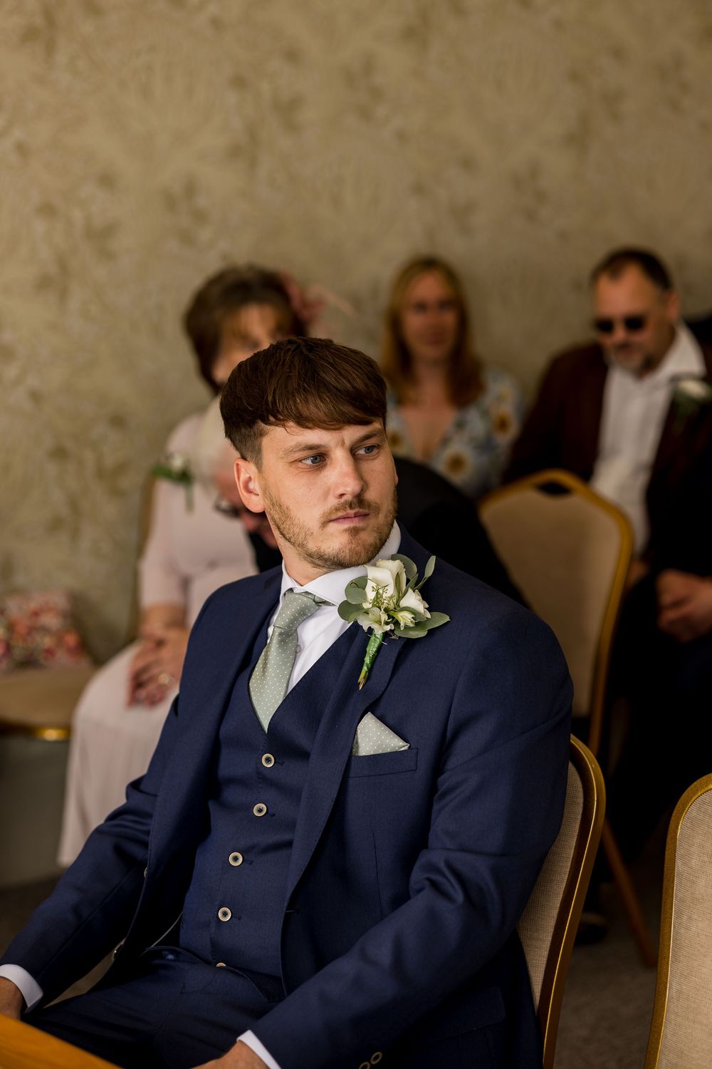 Groom in navy three-piece suit with white boutonniere seated at wedding ceremony with guests behind.