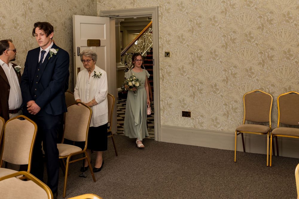Bridesmaid in sage green dress enters wedding ceremony room carrying bouquet, guests standing nearby.