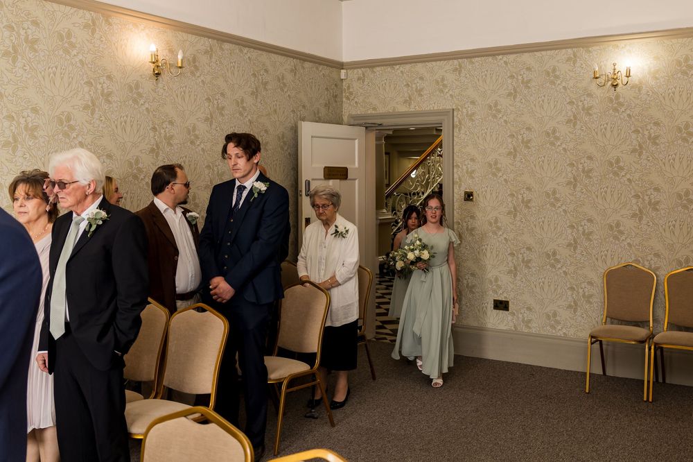 Wedding ceremony guests stand as bridesmaid in sage green dress enters elegant floral wallpapered venue room.