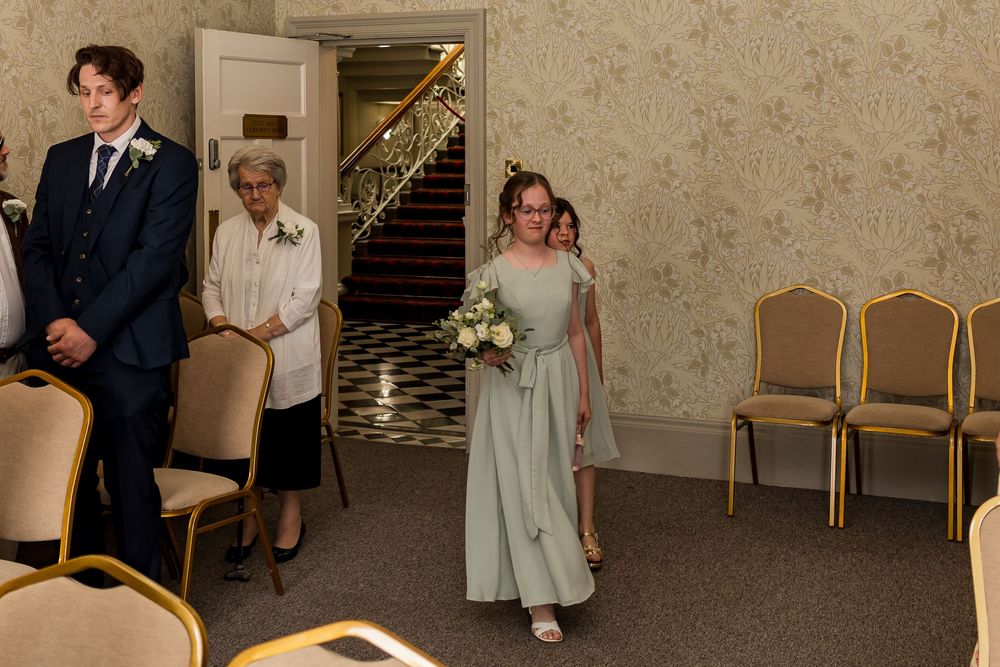 Bridesmaid in sage green dress walks down aisle carrying white bouquet at elegant wedding ceremony.