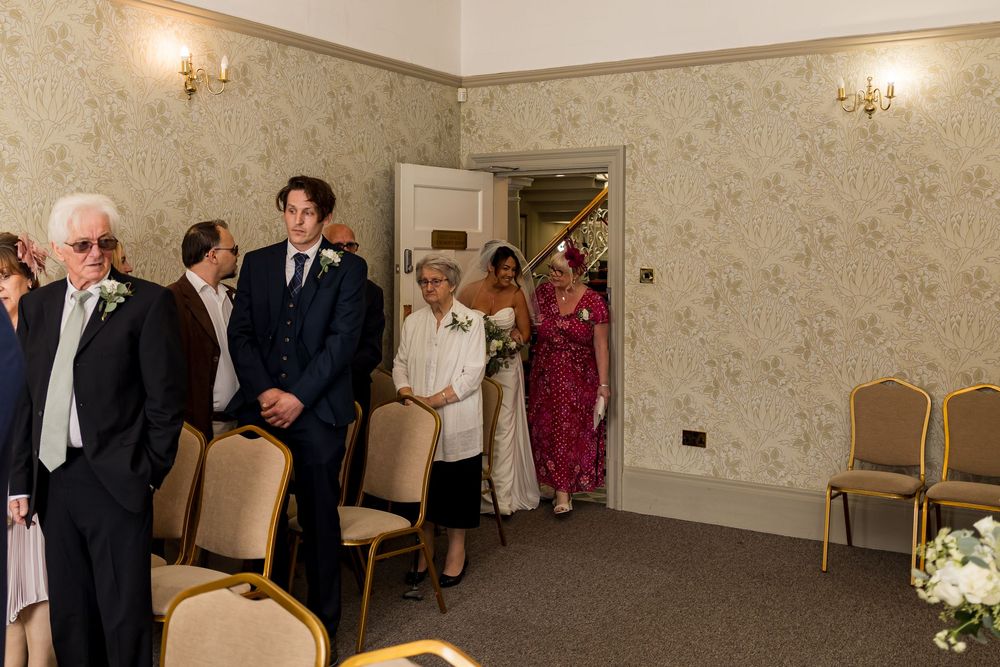 Groom in navy suit awaits bride walking down aisle in elegant wedding ceremony room with floral wallpaper.