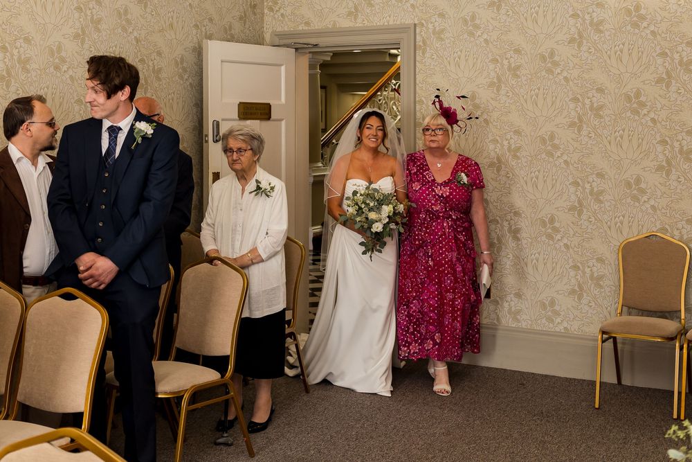 Bride walks down the aisle with family in an elegant wedding venue with gold chairs and floral wallpaper.