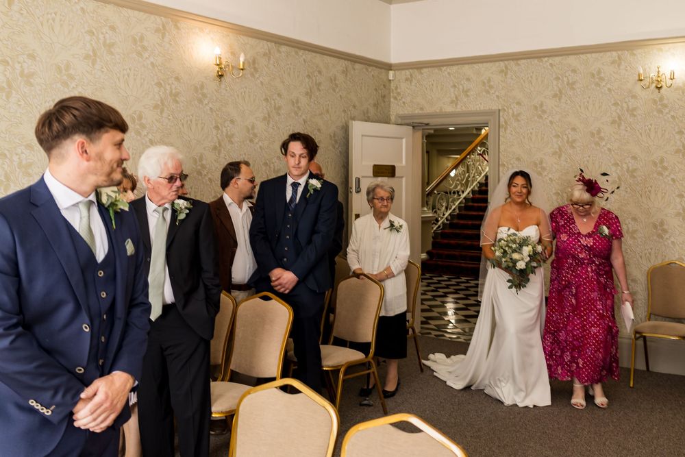 Bride walks down the aisle with family guests standing in an elegant wedding ceremony room.