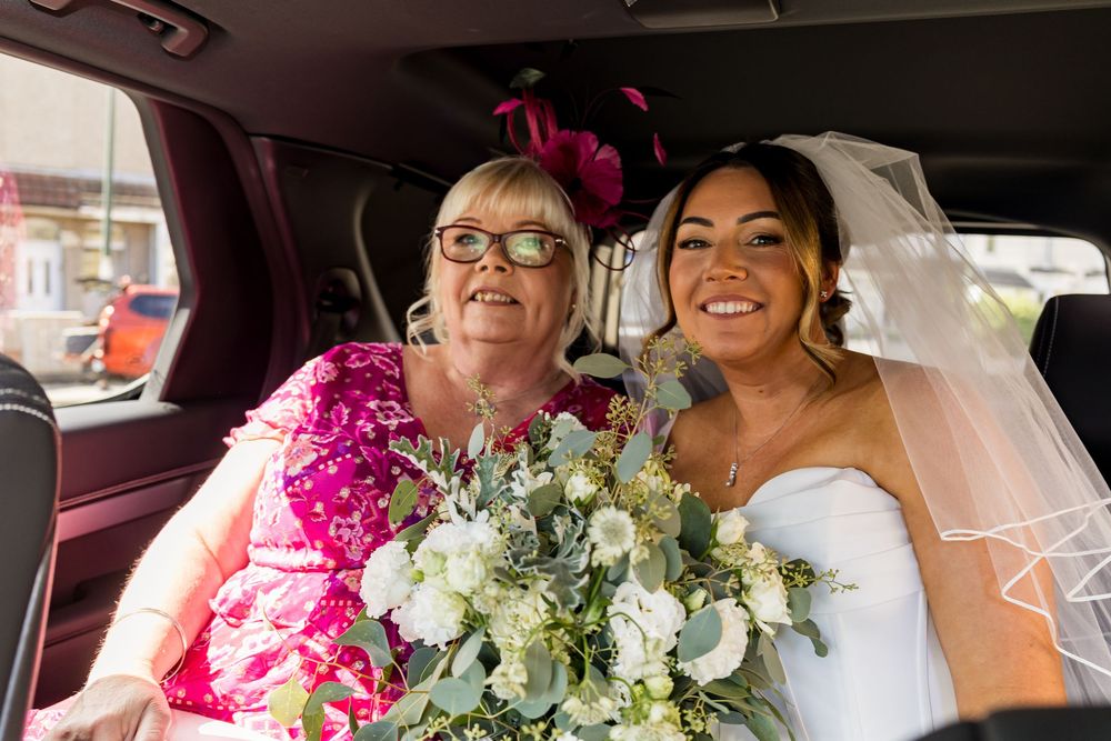 Bride in white strapless gown and veil sits with older woman in pink dress holding bouquet in wedding car.