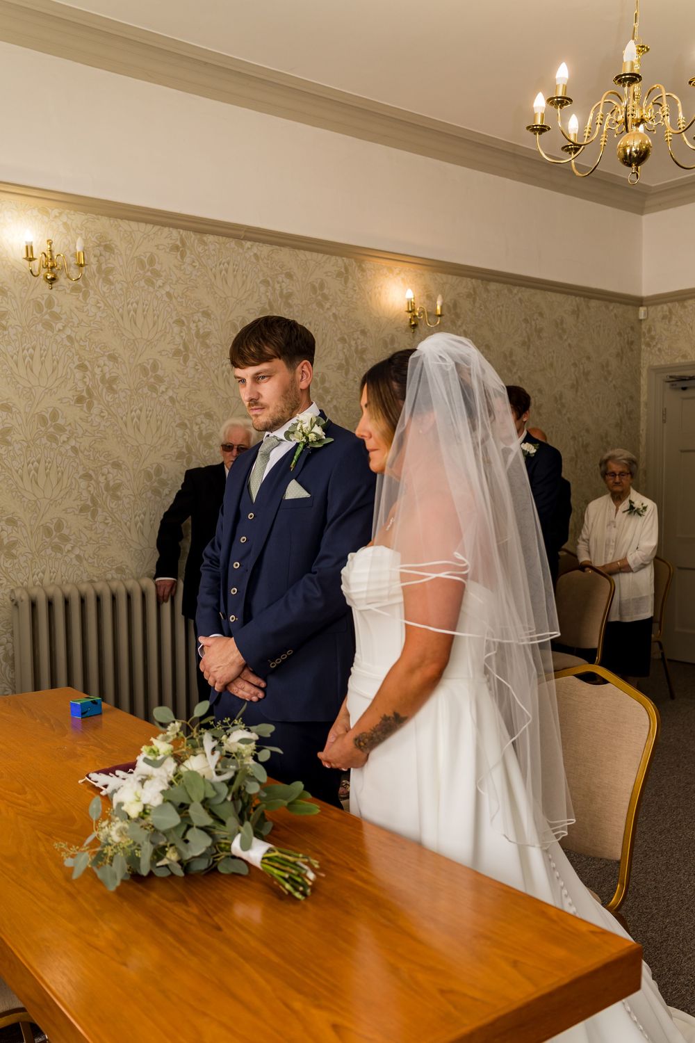Bride and groom stand at ceremony table during indoor wedding service in decorated room.