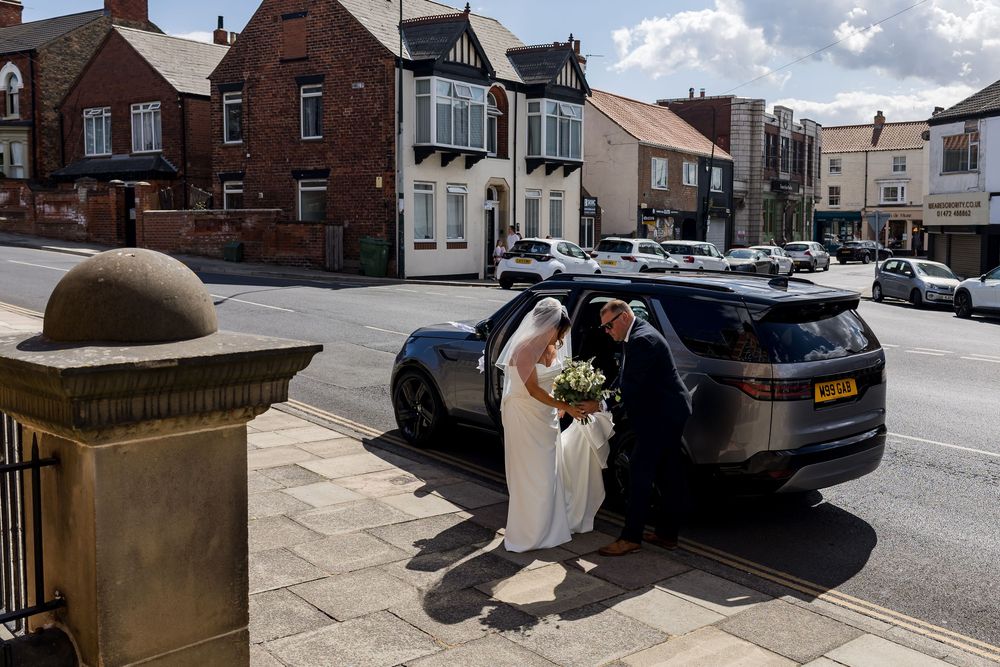 Bride in white gown assisted into a dark Range Rover on a sunny UK high street on her wedding day.