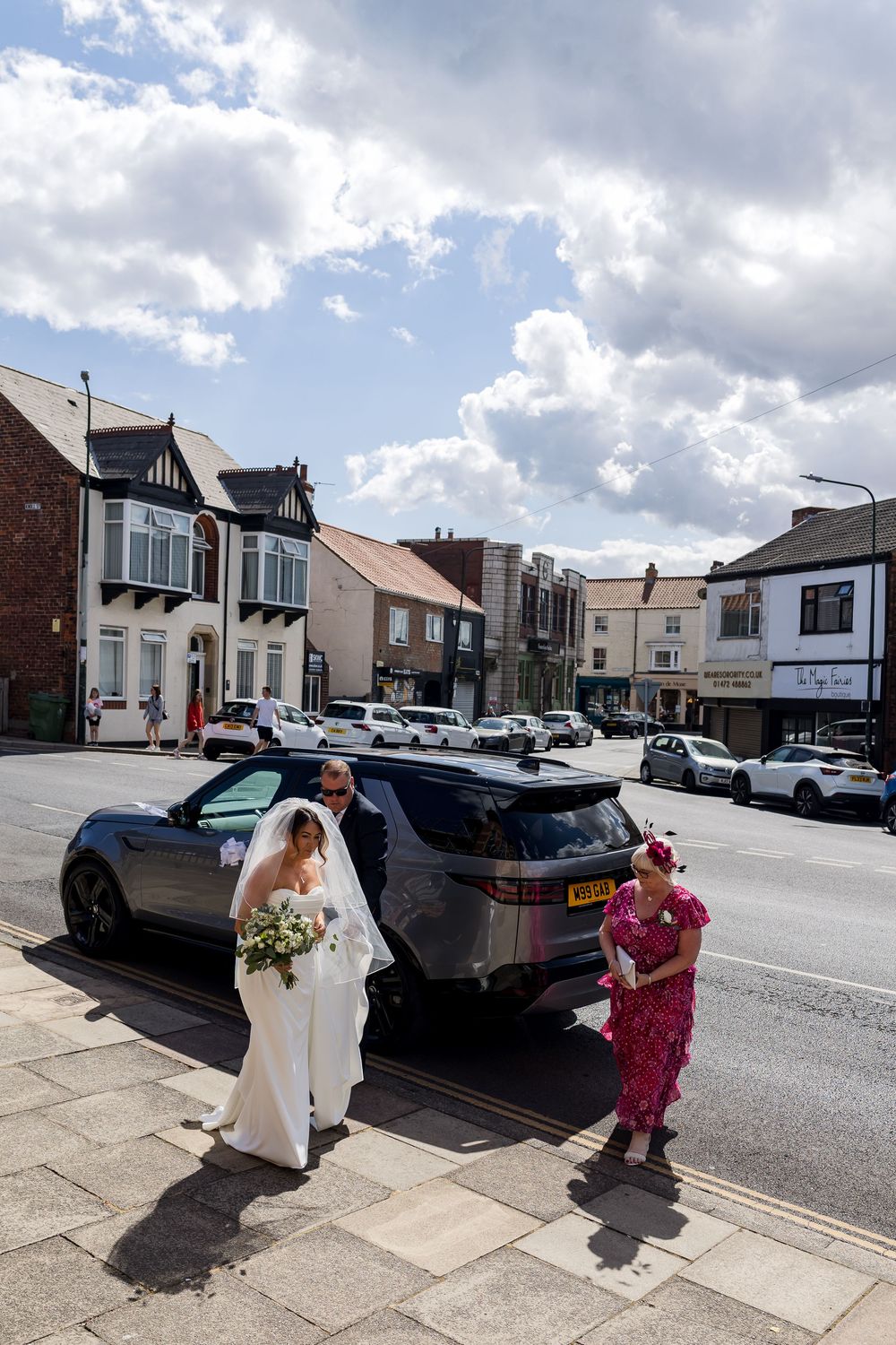 Bride in white gown and veil walks with groom near a Land Rover on a sunny UK high street.