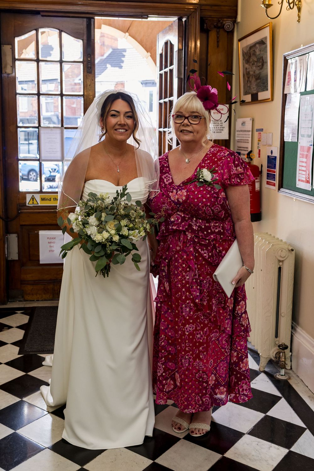 Bride in white gown holding bouquet poses with older woman in red floral dress inside elegant venue.