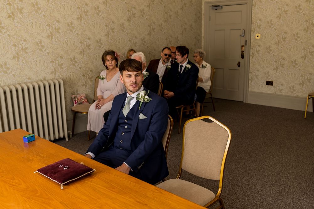 Groom in navy suit sits at wooden table during civil wedding ceremony, guests seated behind in elegant room.
