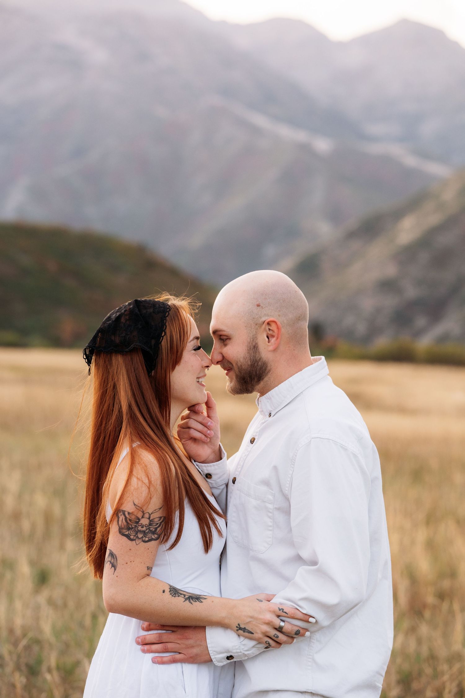 A tattooed couple in white embraces in a golden field with mountain backdrop in a romantic outdoor portrait.