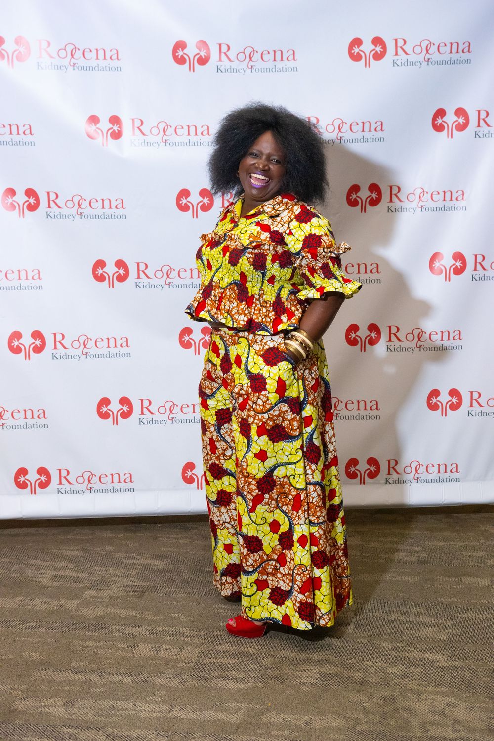 Woman in vibrant African print dress poses at Roeena Kidney Foundation event backdrop.