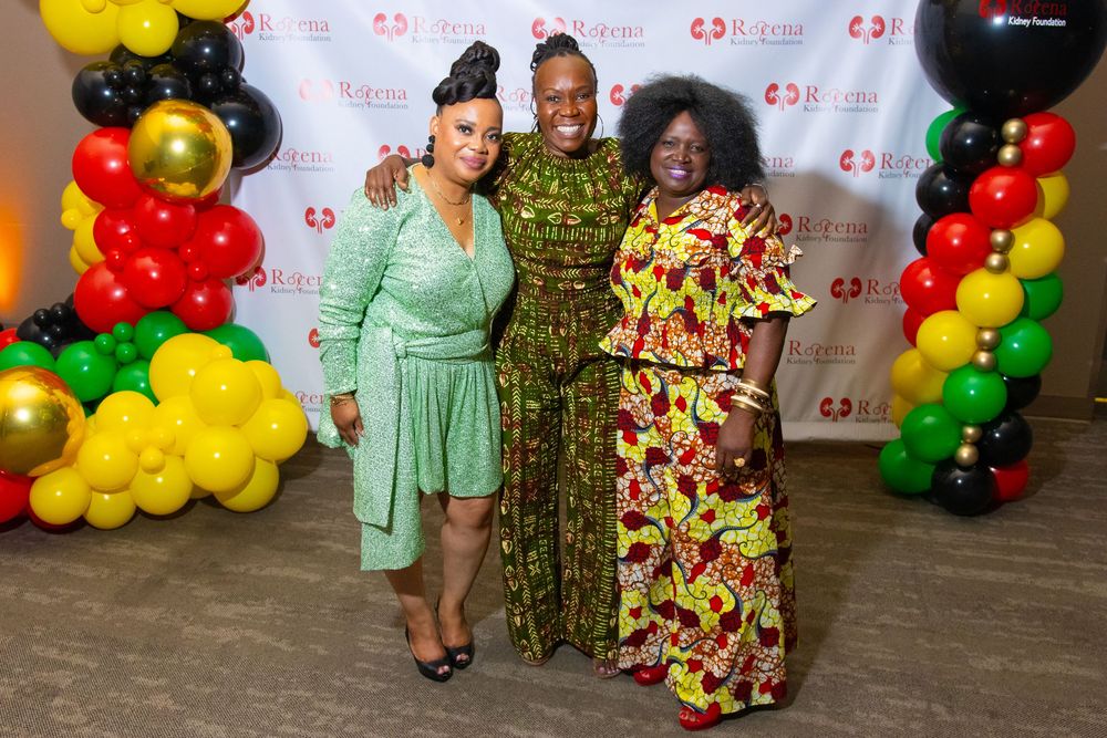 Three women in colorful African print dresses pose together at a festive event with red, gold, and green balloon decor.