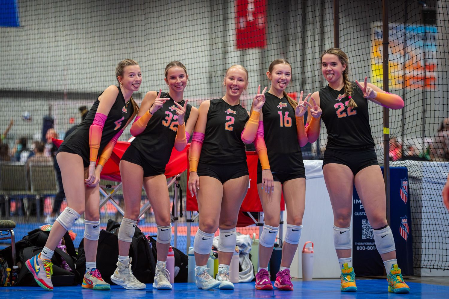 Five female volleyball players in black uniforms smile and pose together on an indoor court during a tournament.
