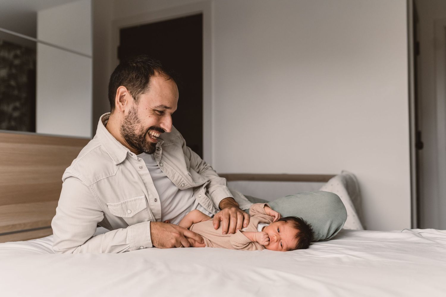 A father tenderly holds his newborn baby near a sunlit window with pink flowers in the foreground.