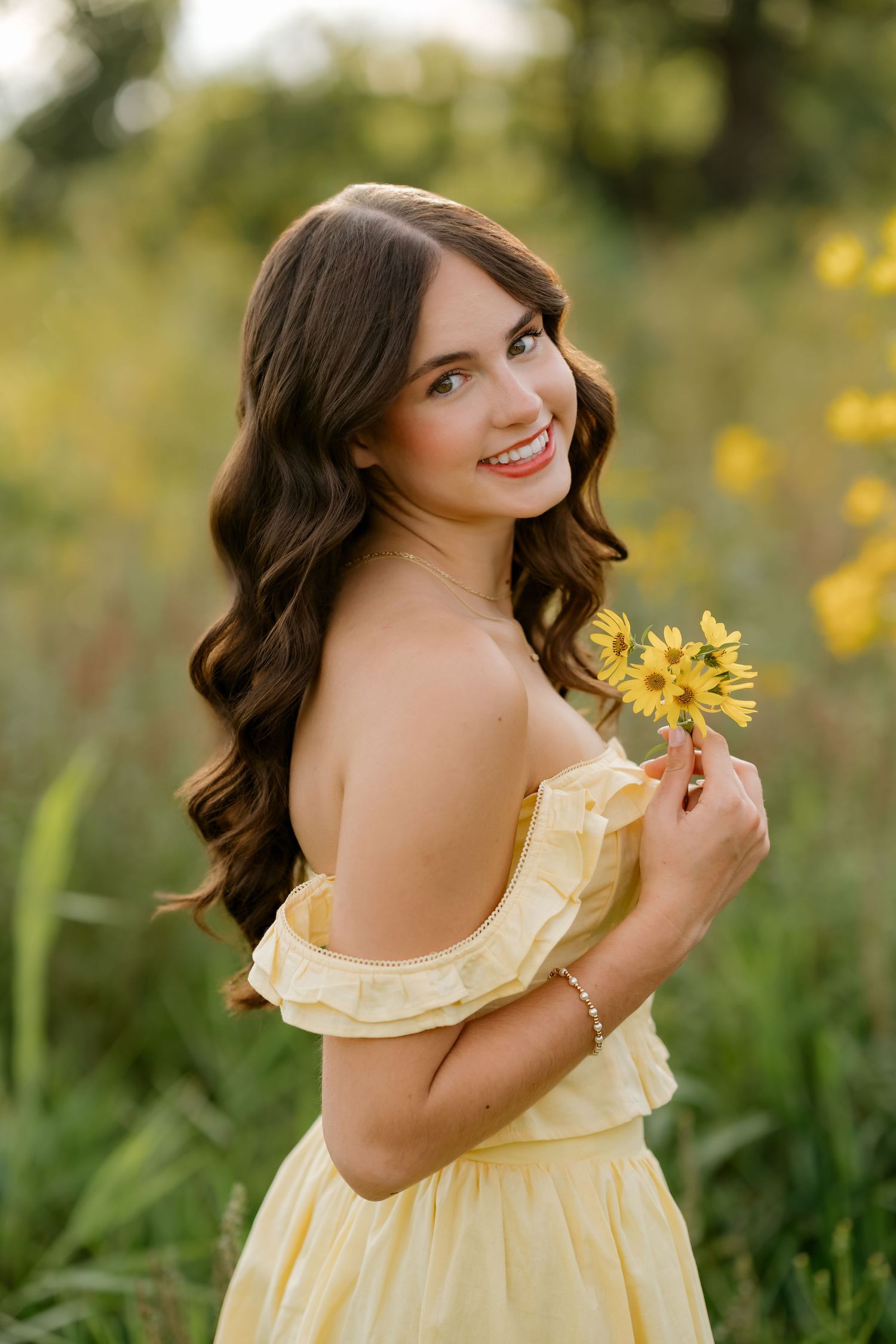 Young woman in yellow off-shoulder dress holding sunflowers, smiling in a lush green field during golden hour.