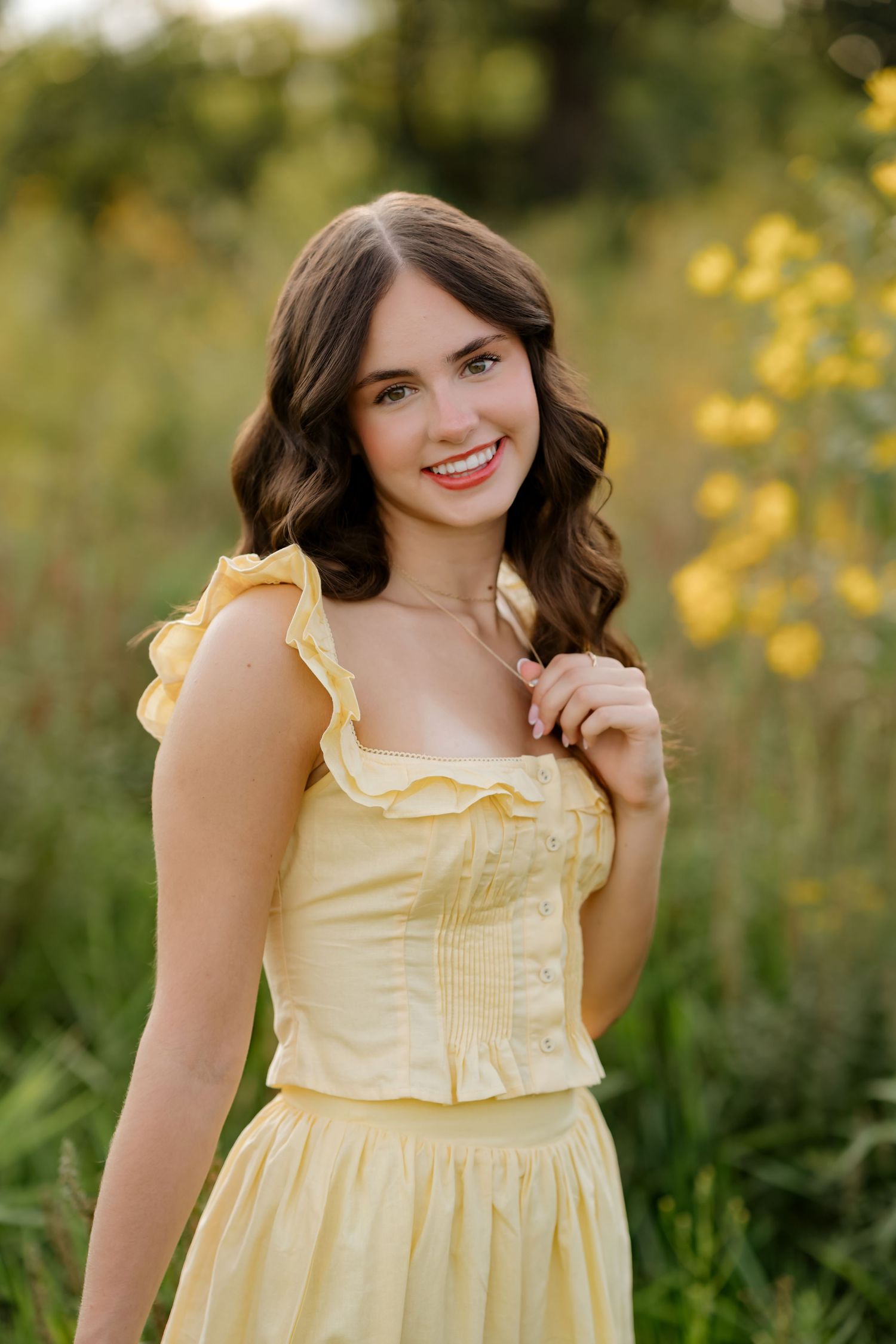 Young woman in yellow ruffled dress smiling outdoors among green grass and yellow wildflowers.