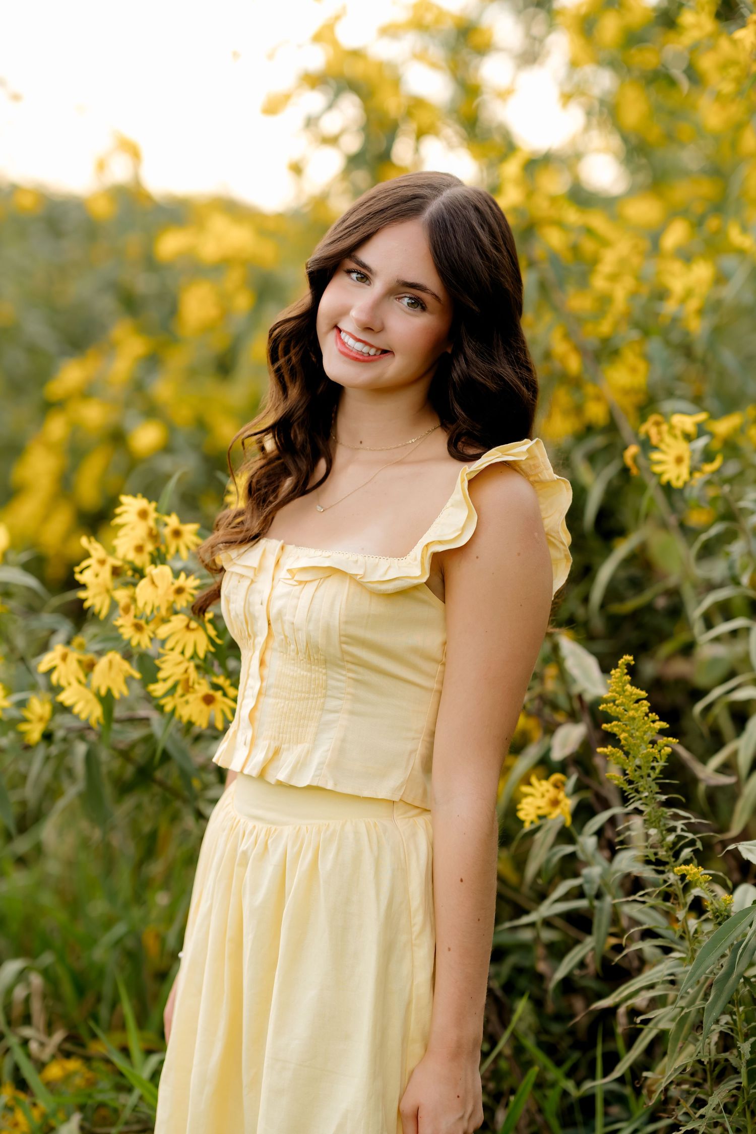 Young woman in yellow ruffled dress smiling among bright yellow wildflowers in a lush green outdoor setting.