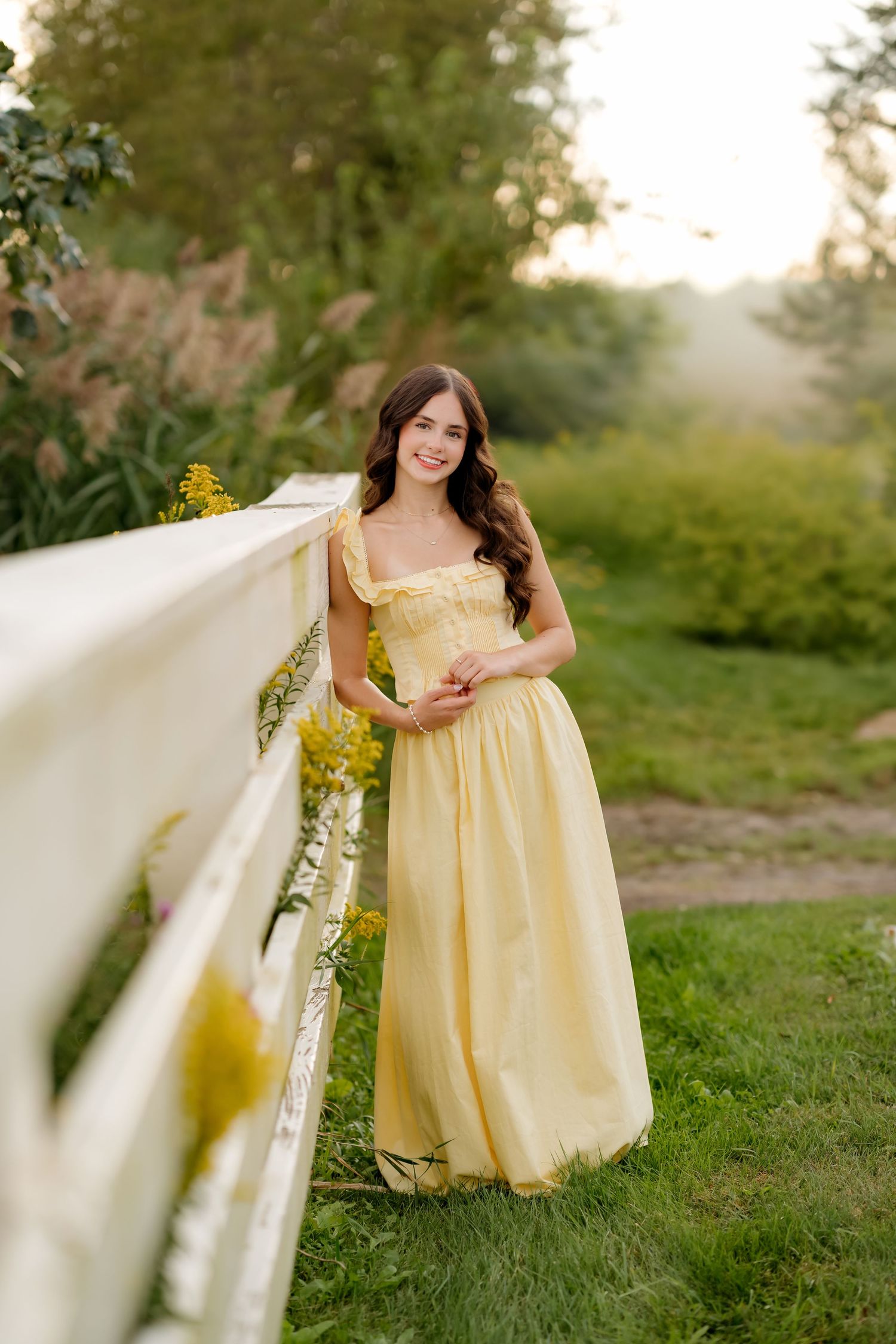 Young woman in yellow maxi dress leaning on white fence surrounded by lush greenery during golden hour.