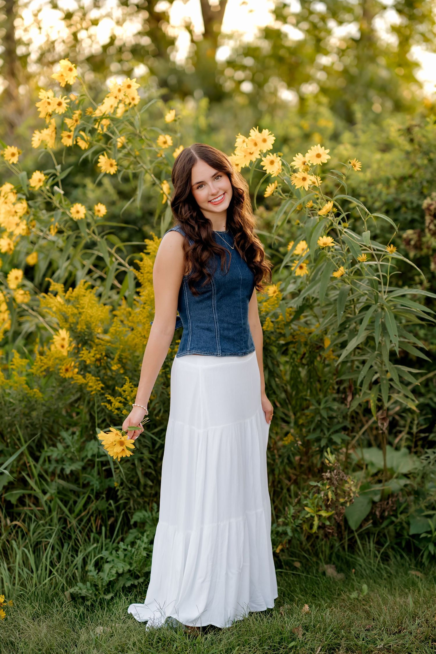 Young woman in denim top and white maxi skirt holding yellow flowers while standing in a sunny garden.
