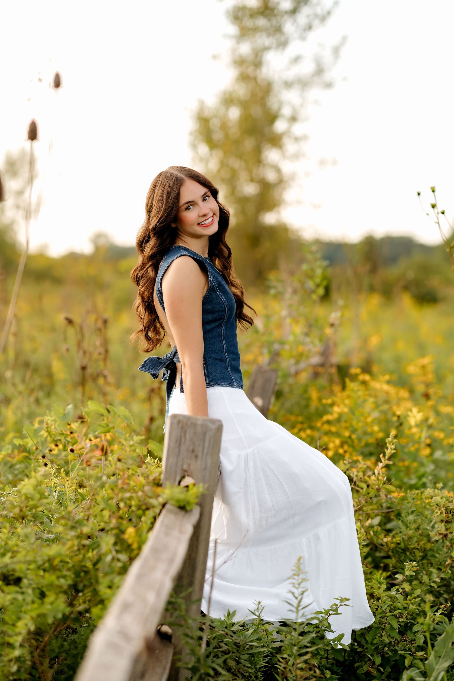 Young woman in denim top and white maxi skirt smiling by rustic fence in sunny wildflower meadow.