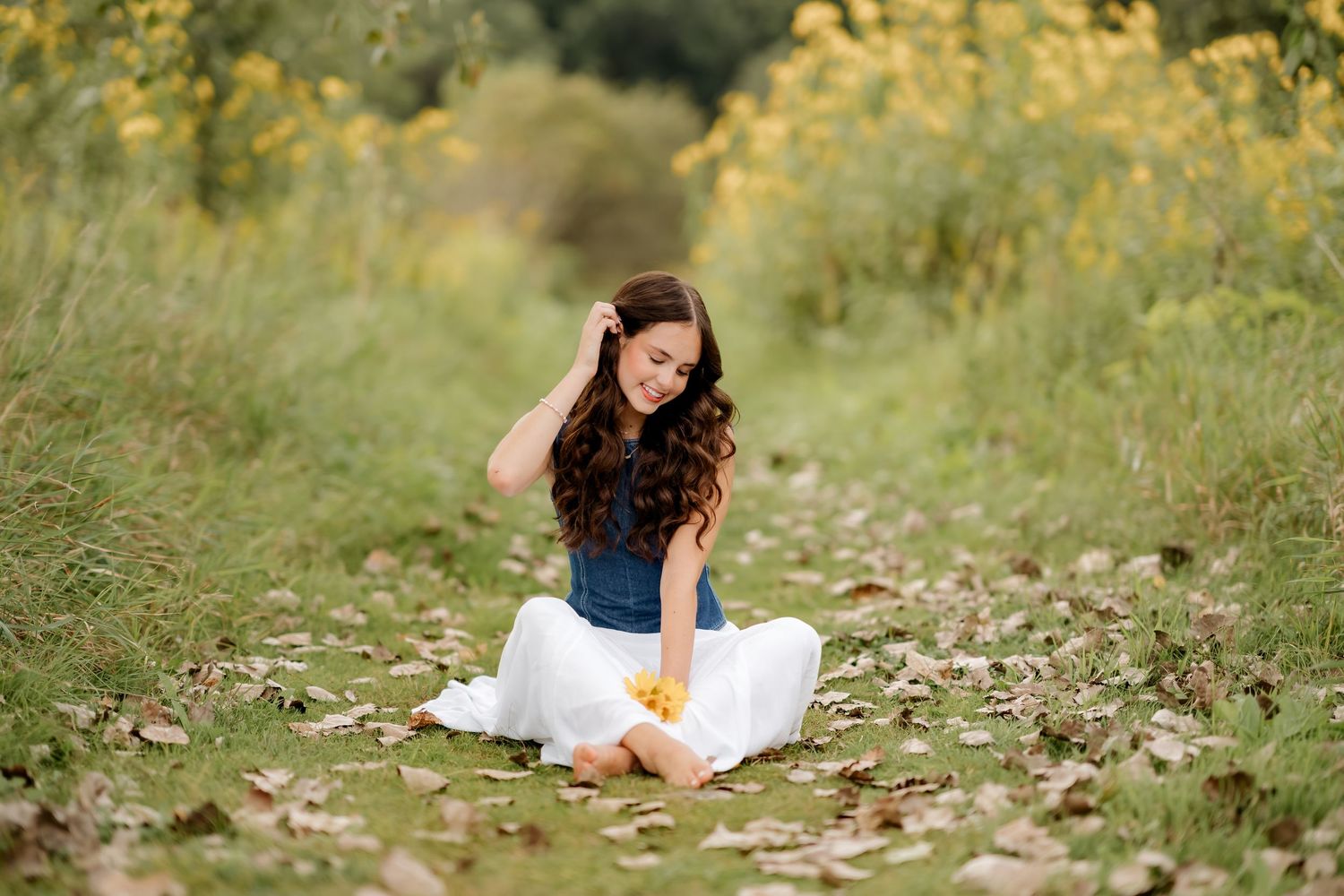 Young woman in white skirt and blue top sitting on a leaf-covered path surrounded by yellow wildflowers.