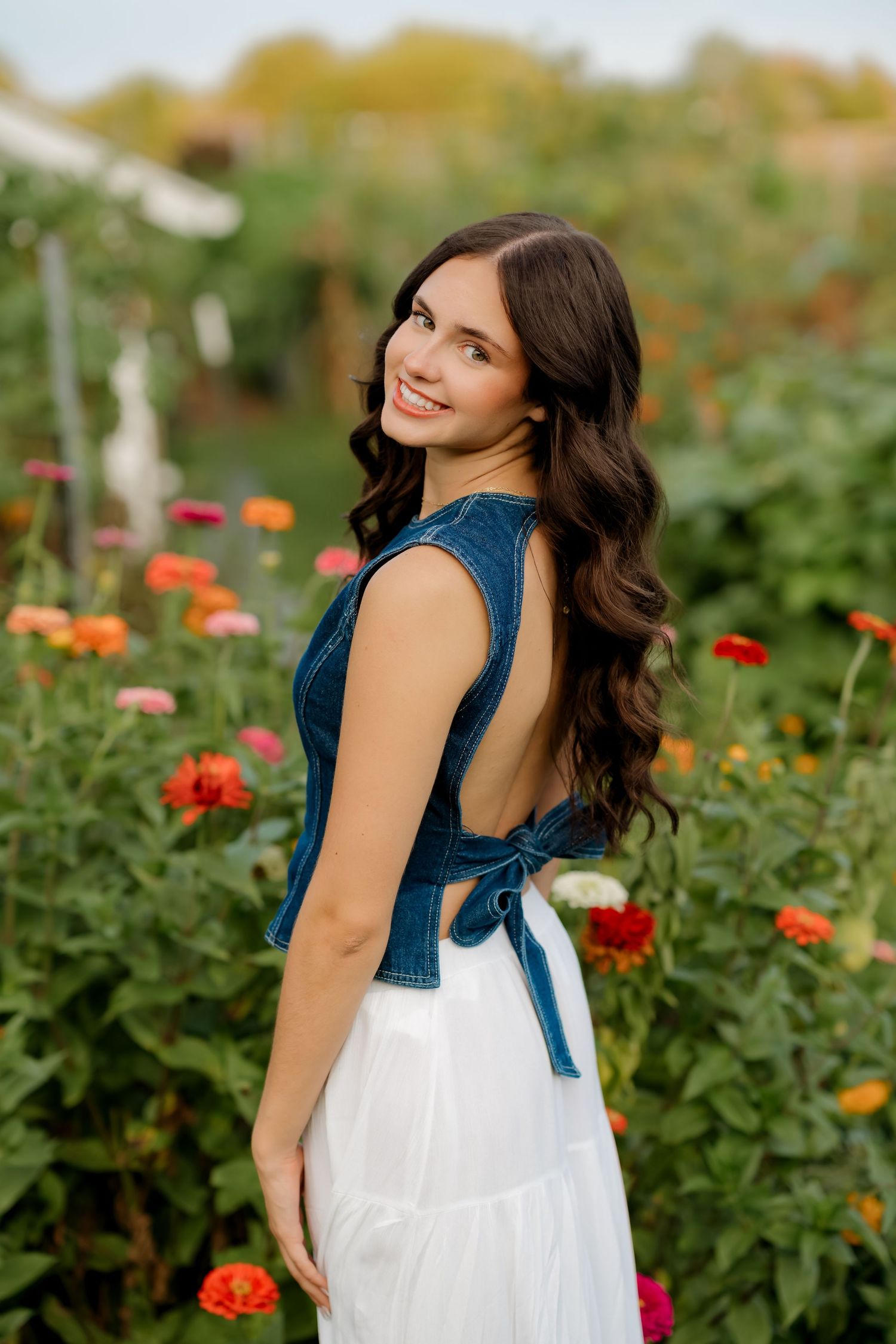 Young woman in backless denim crop top and white skirt smiling over shoulder in colorful flower garden.