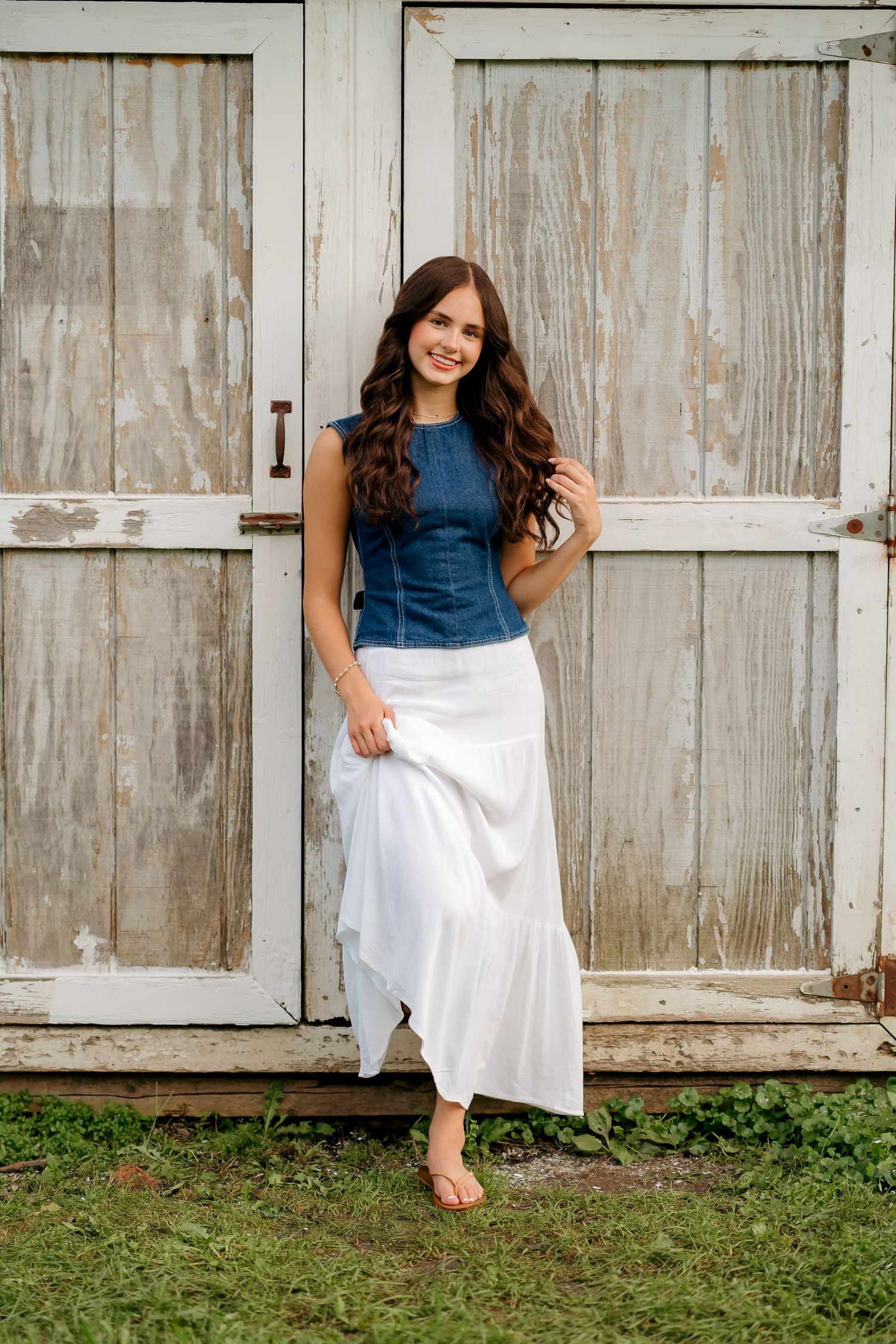 Young woman in denim top and white maxi skirt stands barefoot before rustic wooden doors, smiling.