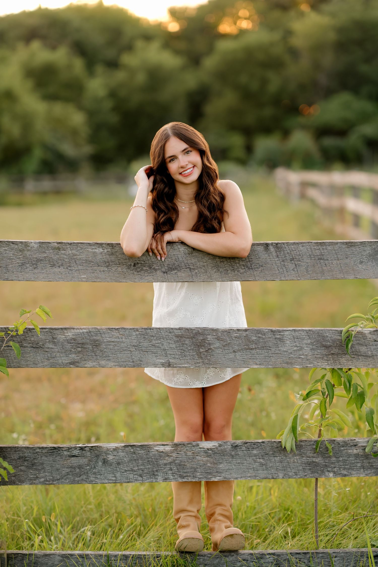 Young woman in white dress and tan boots leaning on rustic wooden fence in golden hour field.