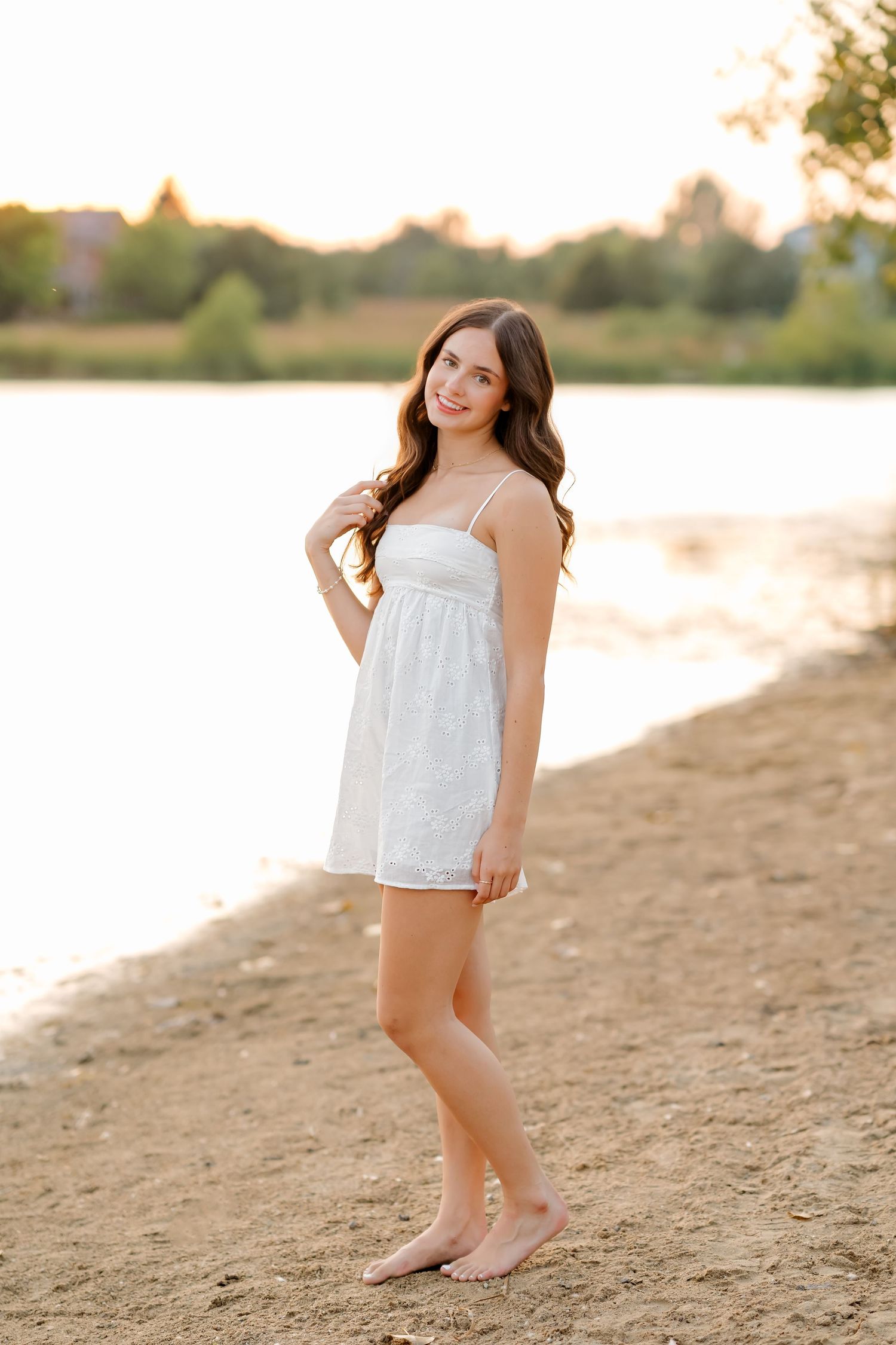 Young woman in white sundress standing barefoot on sandy beach shore at golden sunset hour.