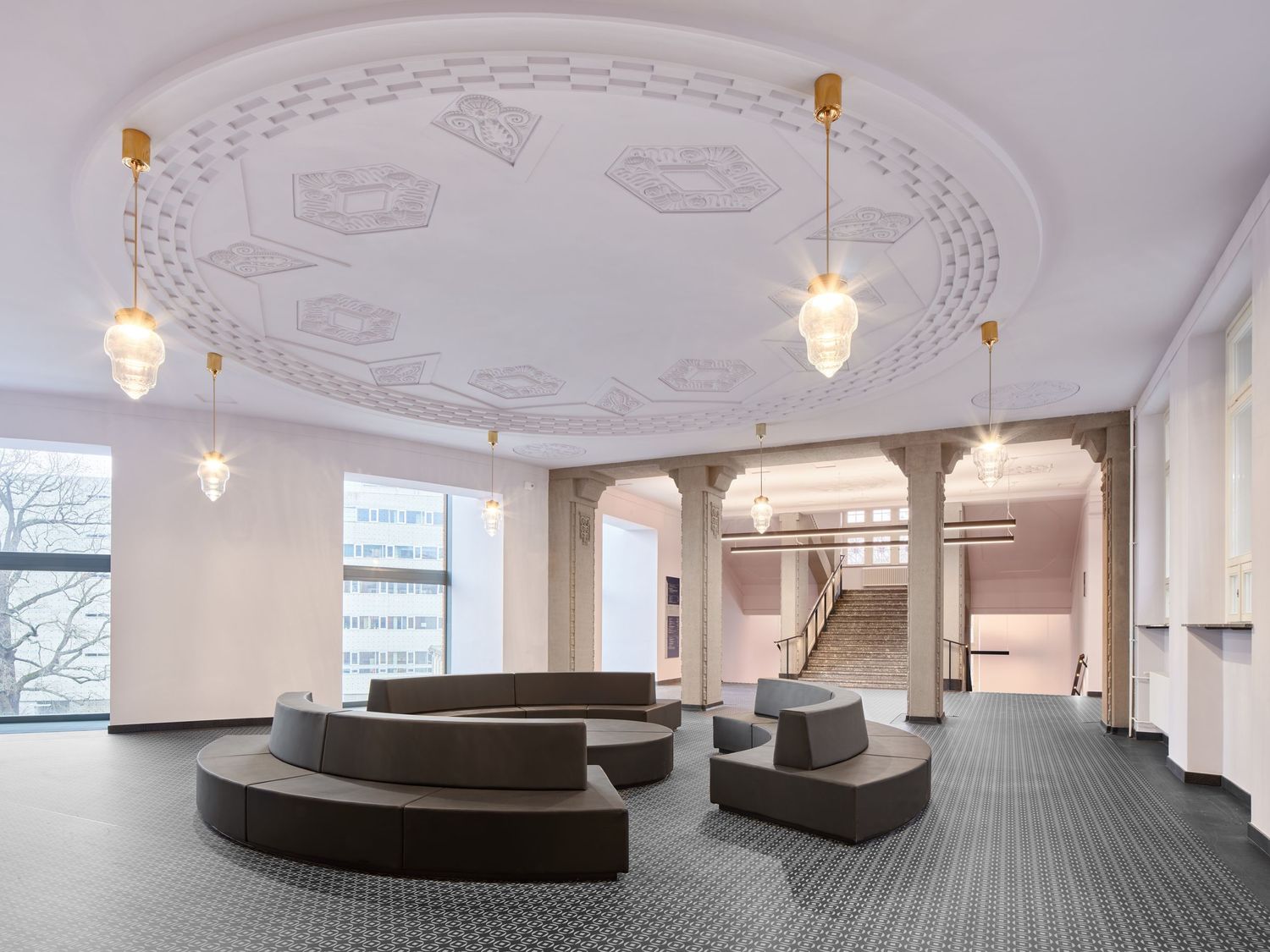 Spacious hall lobby with curved gray sofas, ornate circular ceiling with pendant lights, tall windows and a staircase in the background.