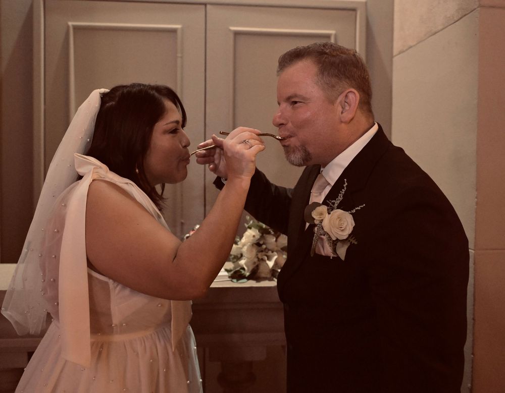 Bride and groom sharing a wedding toast, woman in white gown and veil feeding man in black tuxedo with boutonniere.