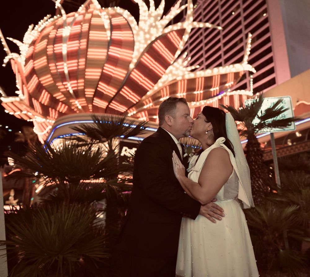 Bride and groom share a romantic kiss on the Las Vegas Strip at night with glowing casino lights behind them.