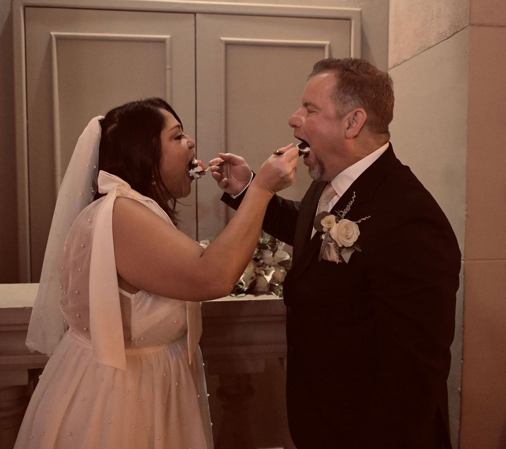 Bride and groom feed each other wedding cake during their reception ceremony indoors.