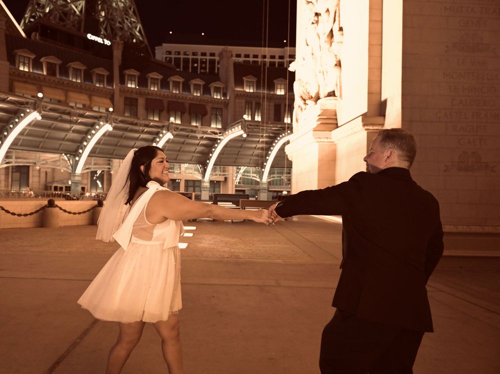Bride and groom dancing joyfully near a fountain outside a grand Las Vegas venue at night.
