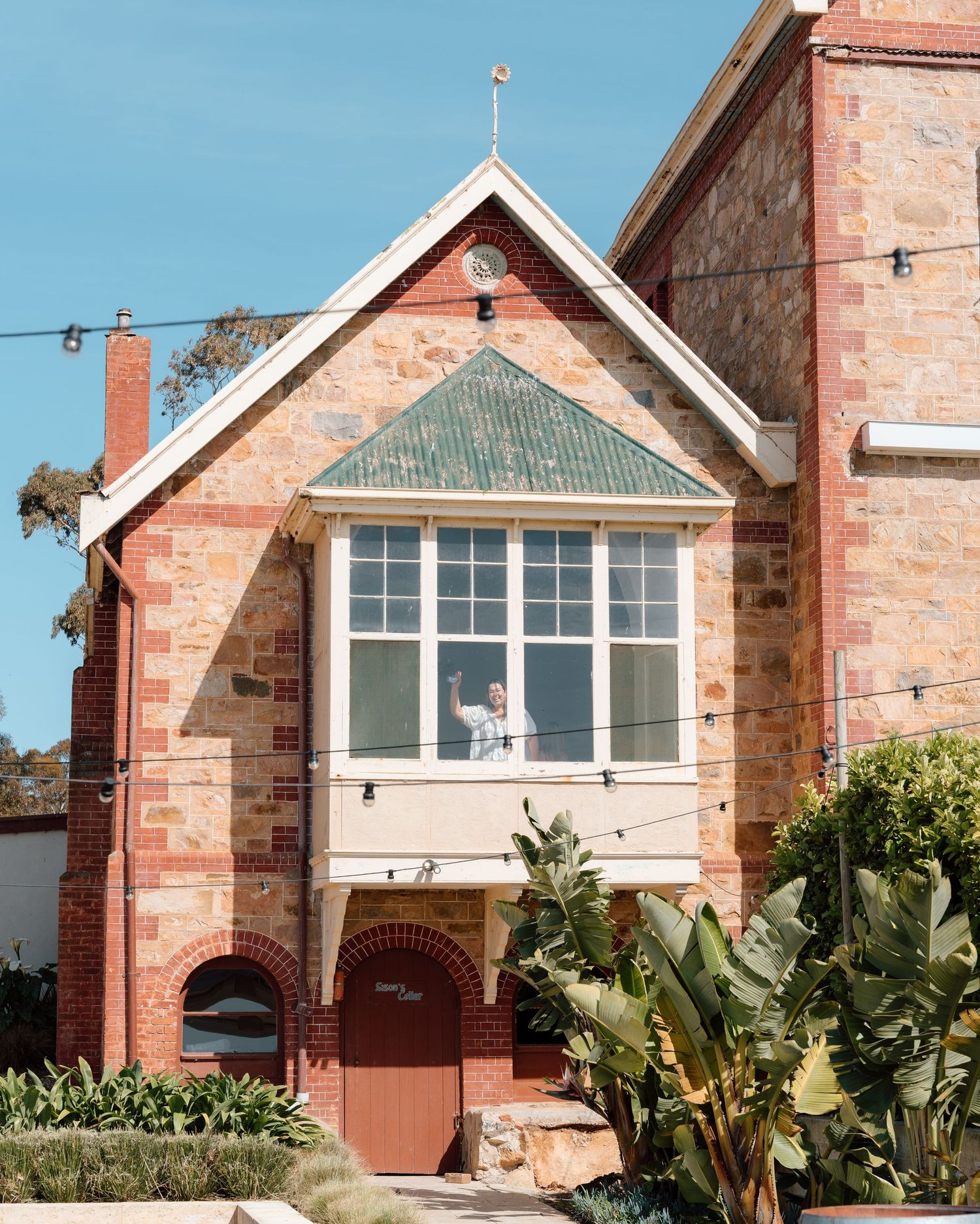 A person waves from a window of a charming red brick building with green roof and arched doorway.
