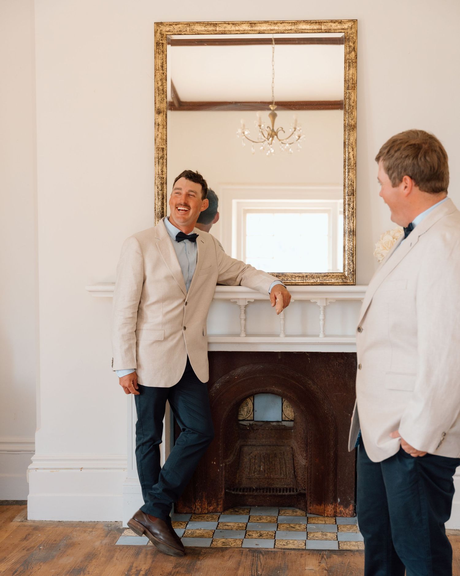 Two men in blazers laughing by an ornate fireplace with gold-framed mirror in an elegant white room.