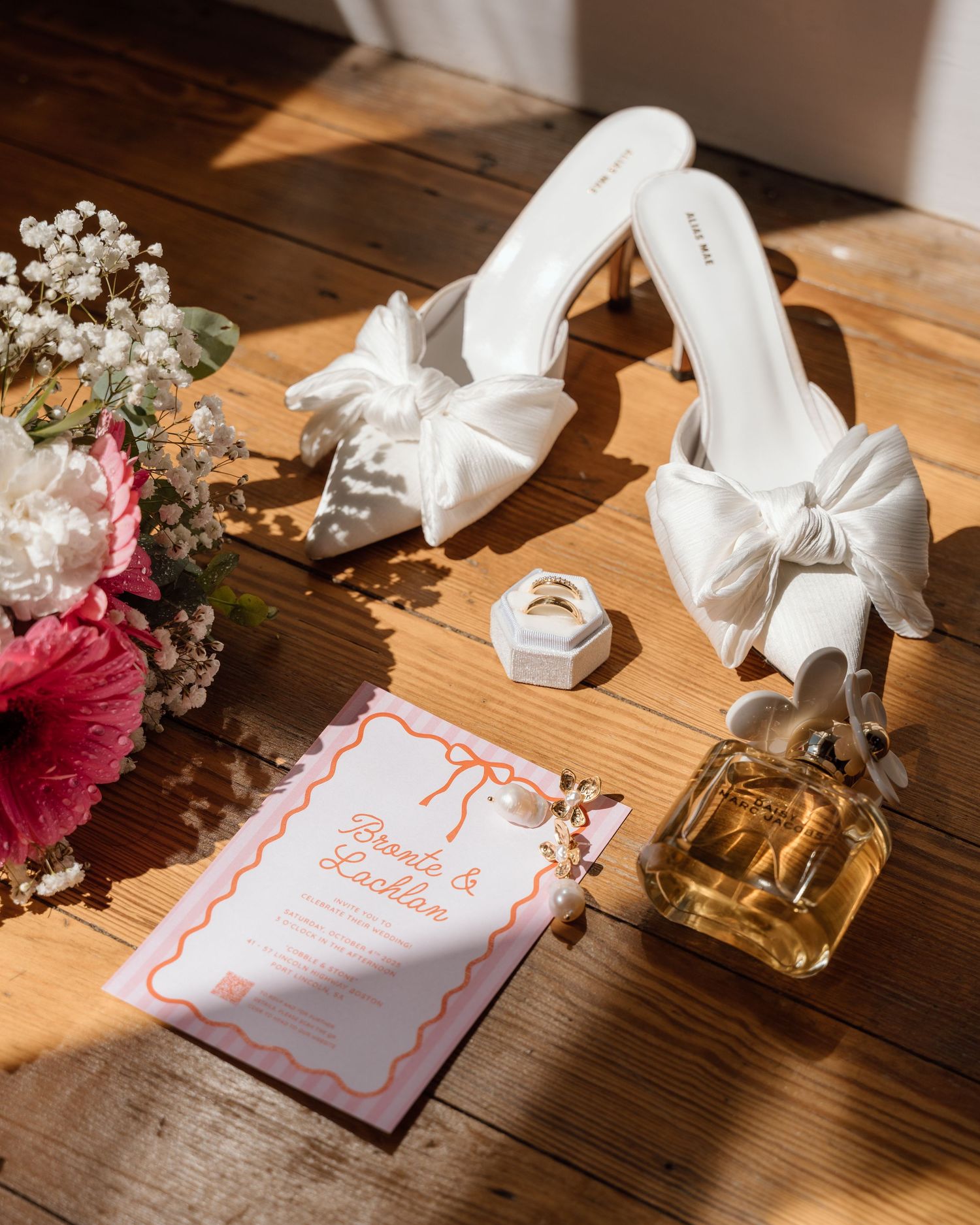 White bow heels, wedding invitation, perfume, ring box, and flowers arranged on a wooden floor in a bridal flat lay.