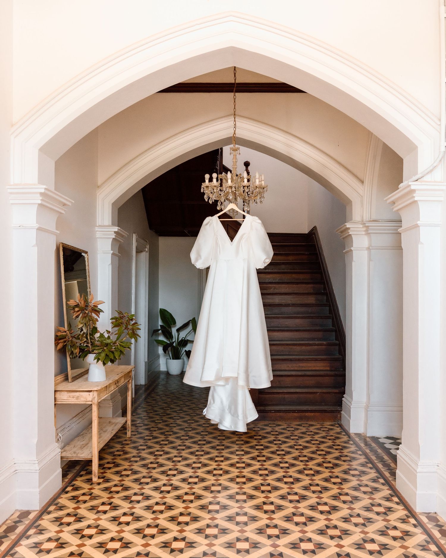 White wedding dress hanging from chandelier in elegant archway with ornate tiled floor and wooden staircase.
