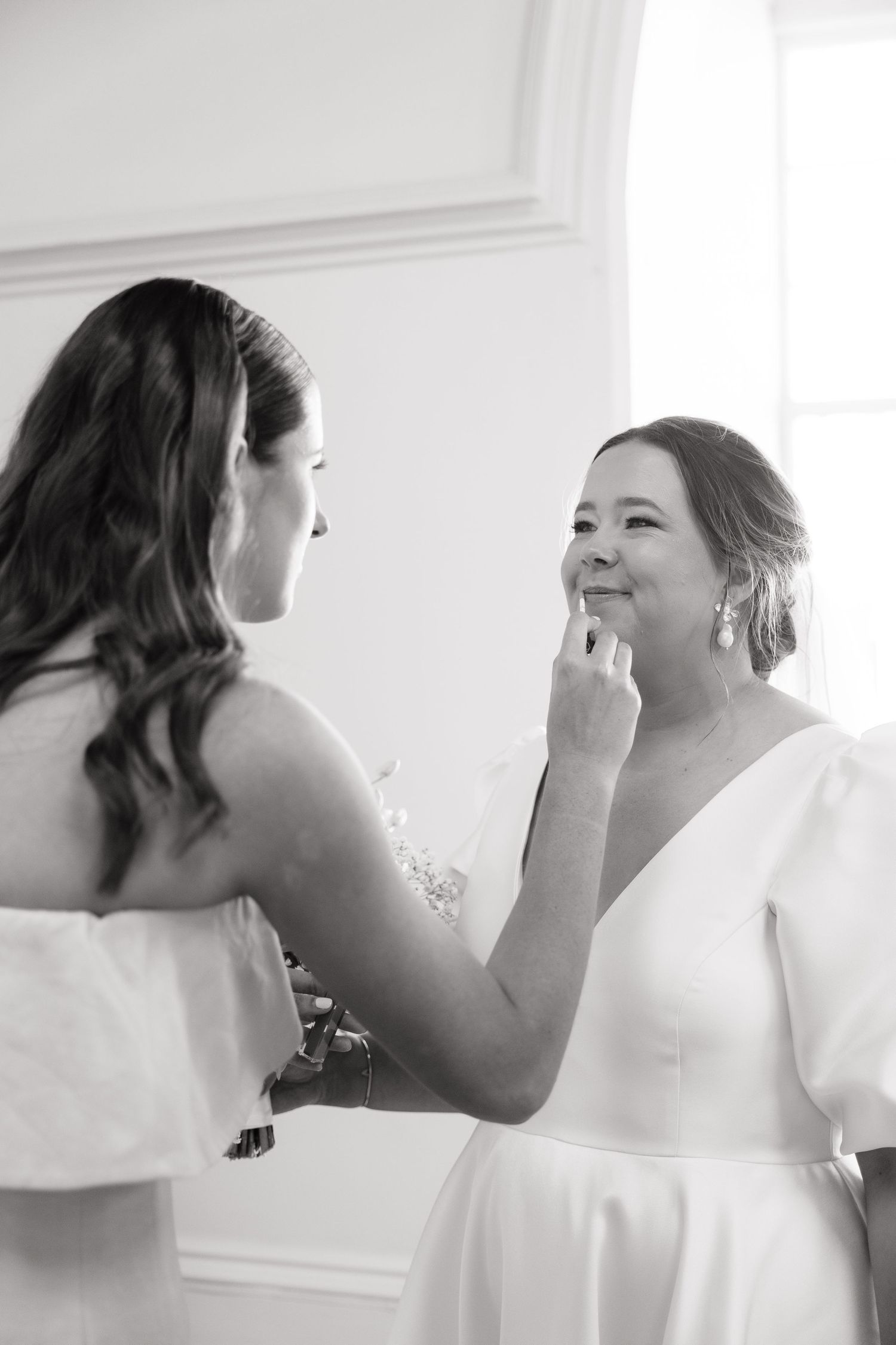 Black and white photo of a bride smiling as her bridesmaid applies makeup during wedding day preparations.