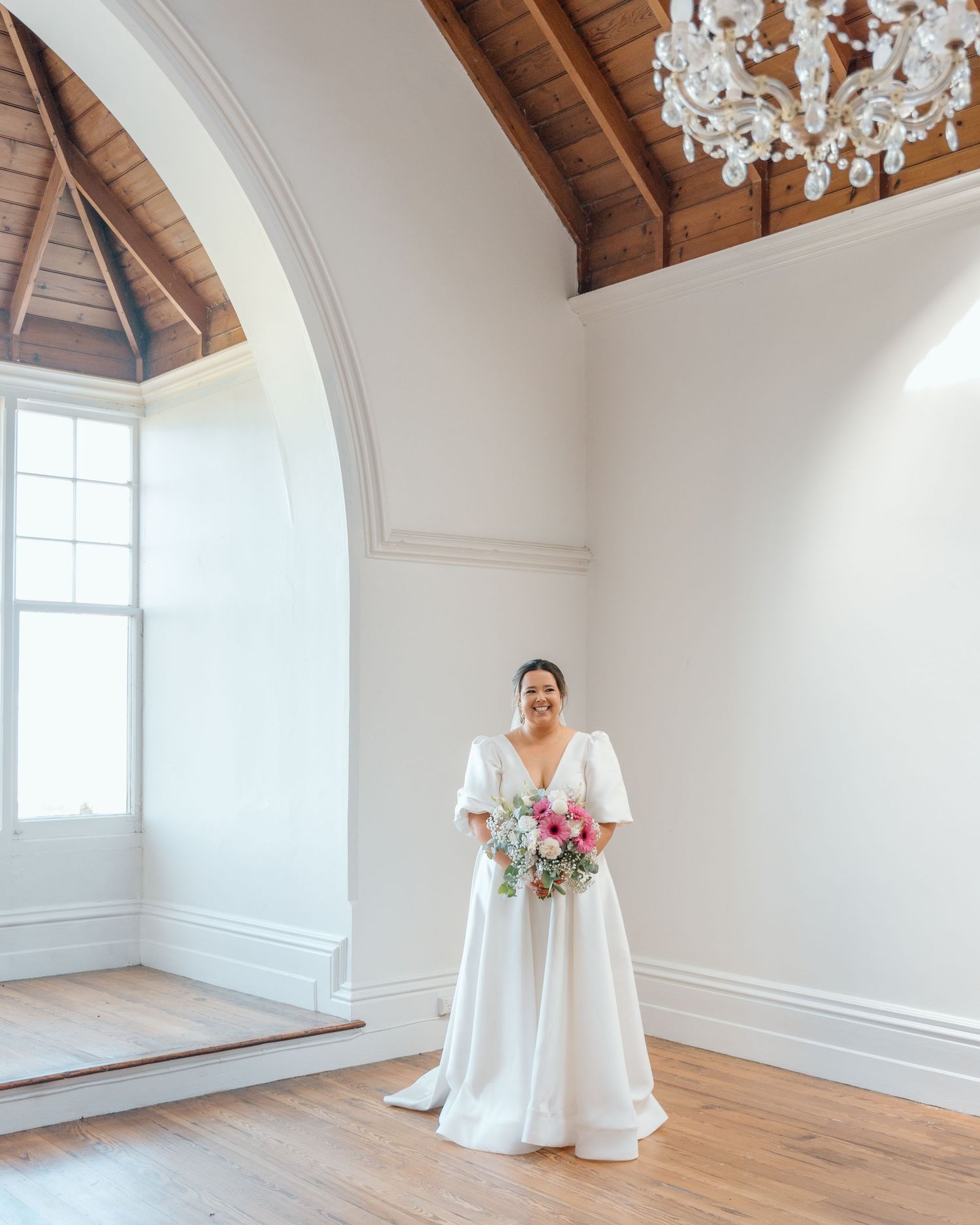 Bride in white flutter-sleeve ball gown holding pink bouquet stands in elegant venue with arched window and chandelier.