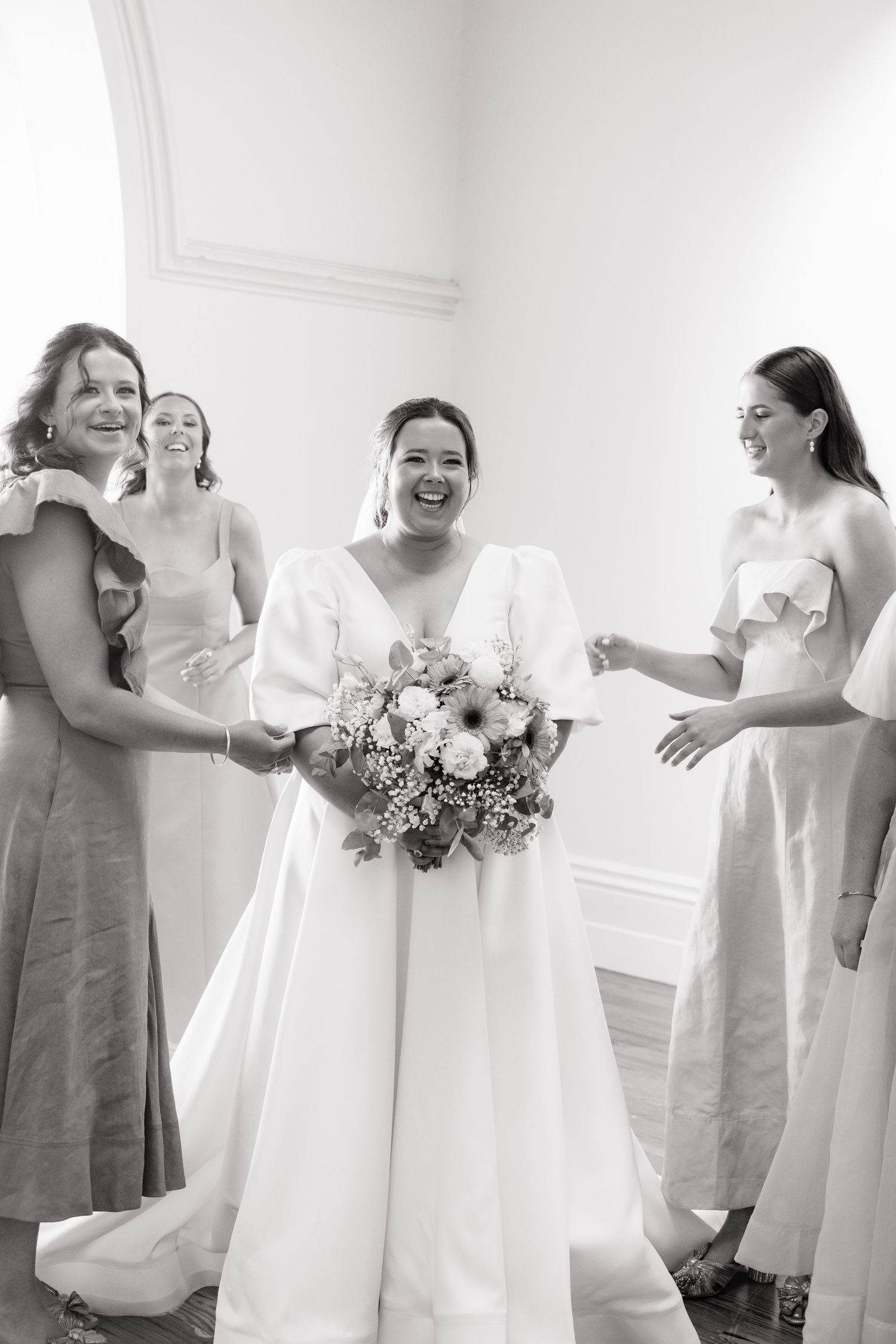 Bride in white gown laughs with bridesmaids holding floral bouquet in black and white wedding photo.