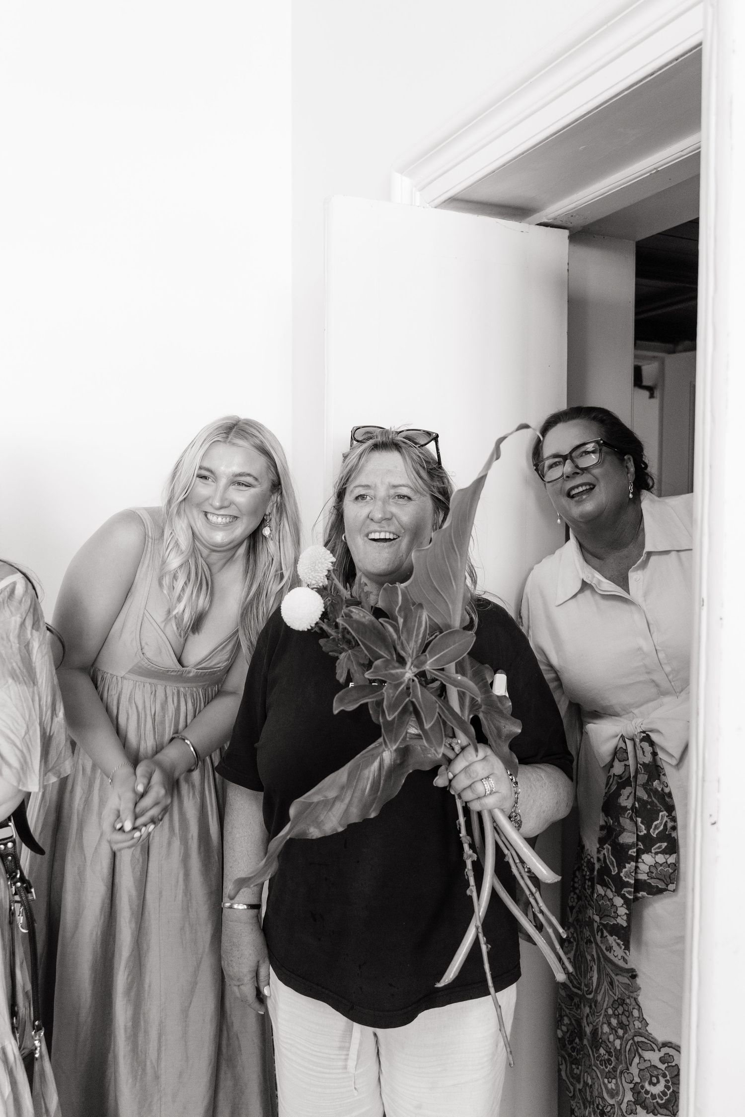 Three women smiling and laughing together at a wedding, one holding a bouquet of flowers, in black and white.