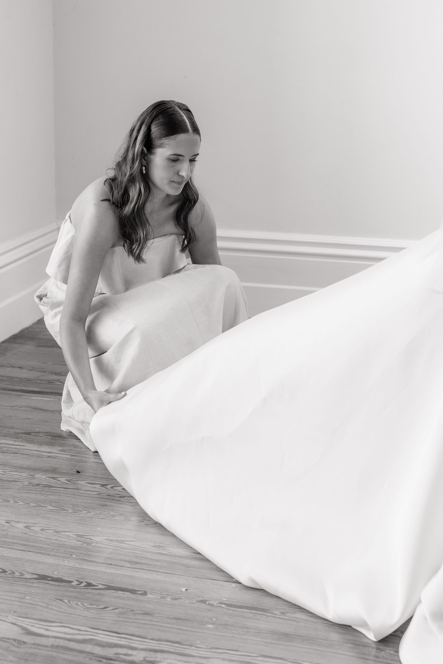 Bride in a strapless wedding gown adjusting her flowing skirt on a hardwood floor in a bright room.