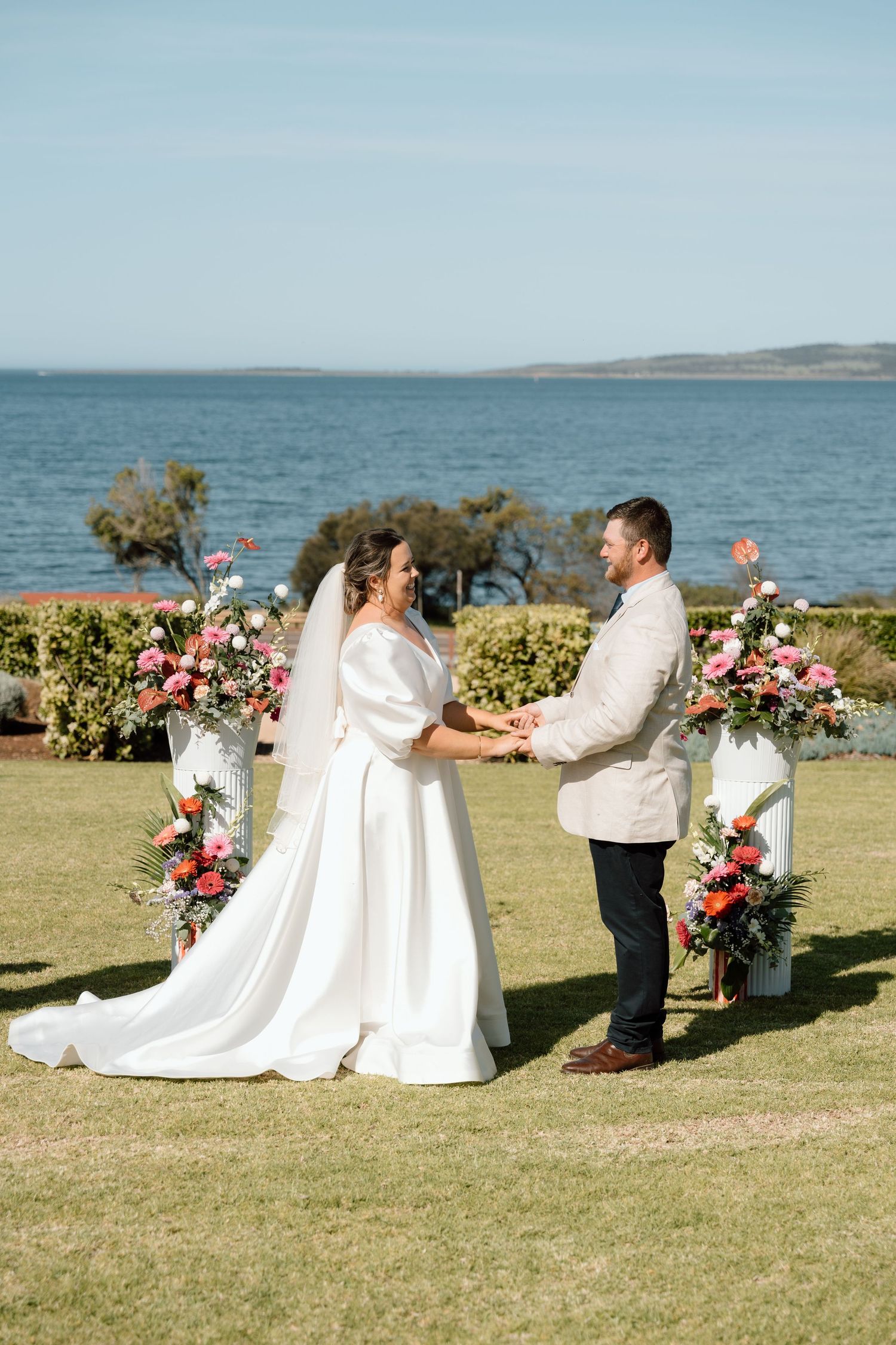 Bride and groom exchange vows outdoors by the ocean, surrounded by vibrant floral arrangements on a sunny day.
