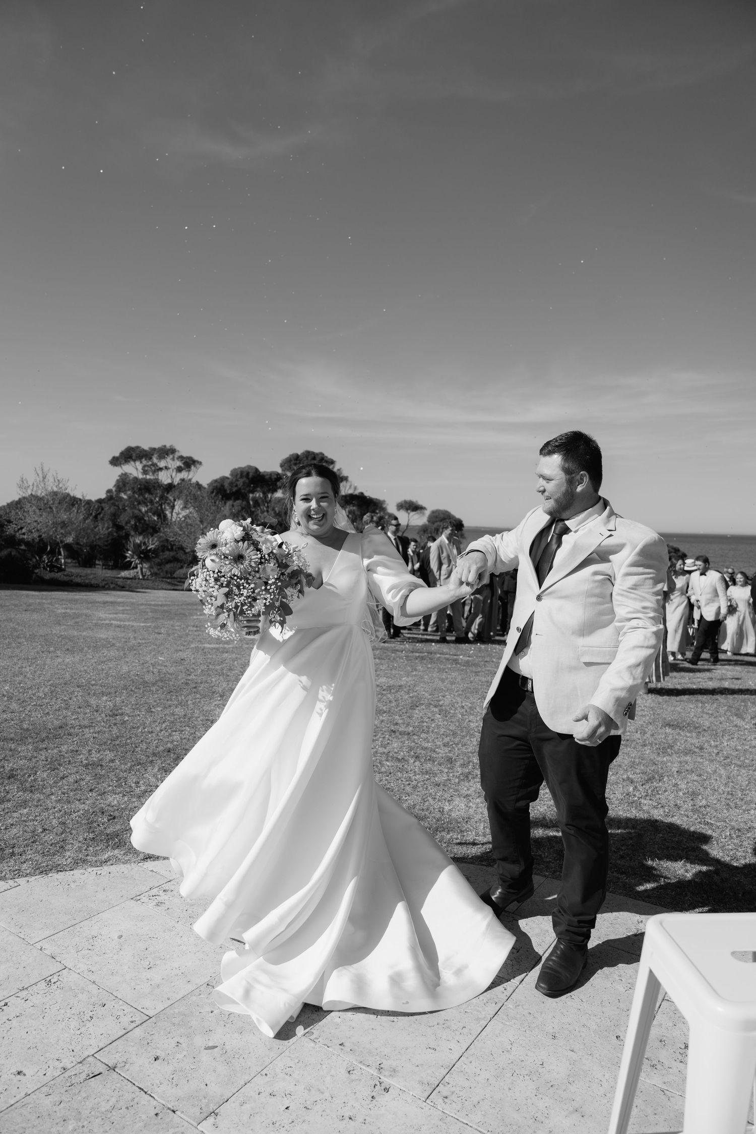 Bride in white gown and groom in light suit dance joyfully outdoors after their wedding ceremony.
