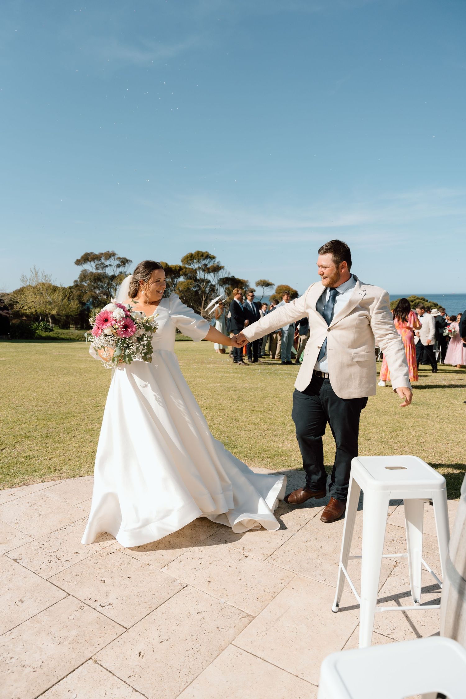 Bride and groom hold hands walking down the aisle at an outdoor coastal wedding ceremony on a sunny day.