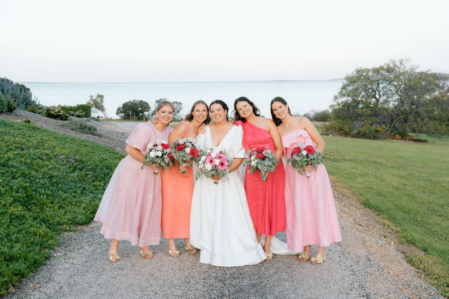 Bride with four bridesmaids in pink and coral dresses holding bouquets, posing outdoors near waterfront.